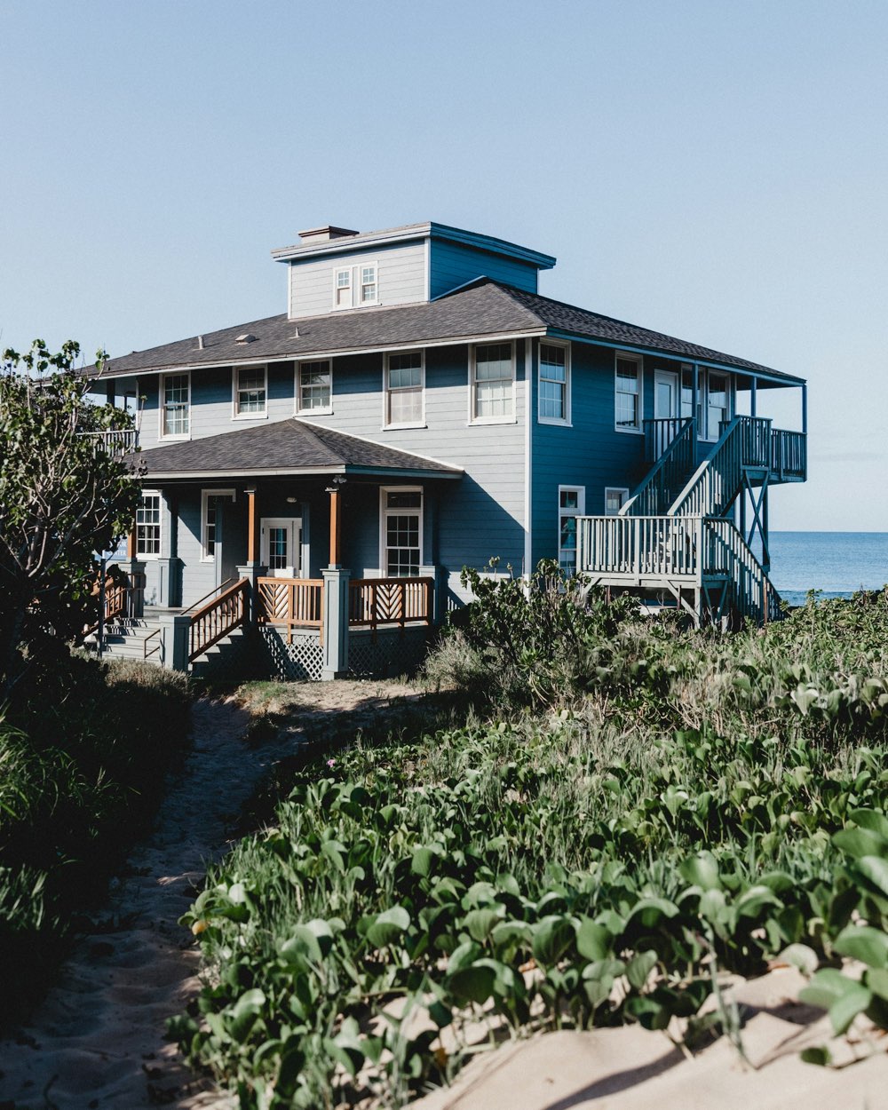 A grey shingled coastal home with wraparound porches, seen from a sandy path through dune grass.