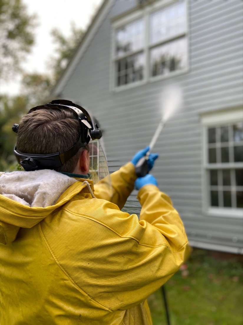 A worker in a yellow jacket pressure washing the clapboard siding of a house.