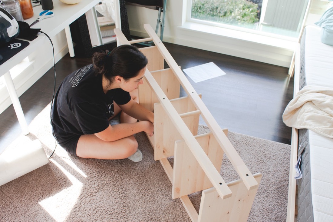 A person kneeling on a bedroom floor assembling a flat-pack wooden furniture frame.